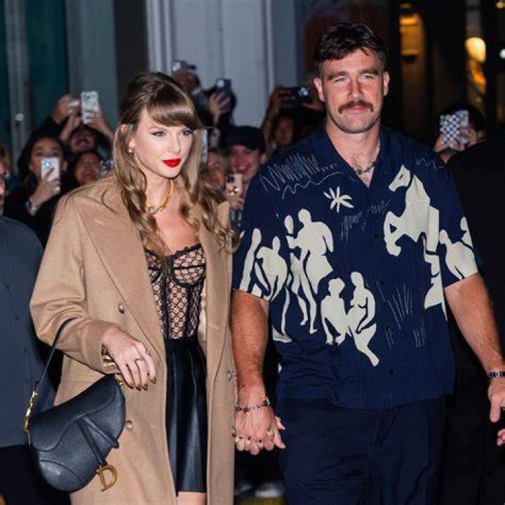 Taylor and Joe walking through a crowd with security around them, keeping their heads angled down, they are holding hands