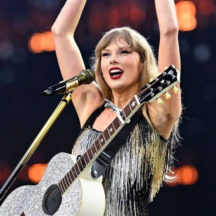 Taylor stood on a high platform singing on her tour, she is wearing a lavender colored coat and is surrounded by purple clouds and lighting